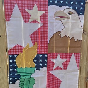 May include: A patriotic garden flag featuring a patchwork design with an American eagle, stars, and the Statue of Liberty's torch. The flag has a red and white checkered pattern and a blue background with white stars. The flag is made of a lightweight material.