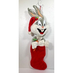 May include: A plush Bugs Bunny character wearing a red Santa hat with a white trim, emerging from a red Christmas stocking. The character has gray and white fur, long ears, a pink nose, and is holding a small orange felt carrot in its hands.