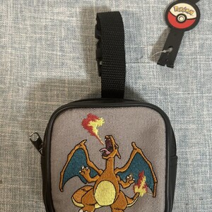 May include: A gray square pouch with a black zipper and strap, featuring an embroidered Charizard design. The pouch has a black border and a Pokemon key tag attached.