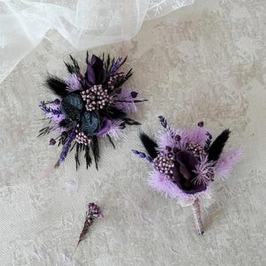 以下が含まれることがあります： 2つの花のブートニアと小さな花の茎。ブートニアは紫と黒のドライフラワーと羽が特徴です。1つのブートニアは、もう一方よりも大きく、手の込んだものです。背景は、白のレース生地のある明るい灰色の表面です。