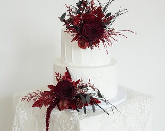 May include: A two-tiered white cake decorated with dark red flowers and greenery. The cake is on a white cake stand and sits on a white lace tablecloth. The flowers include roses and other textured elements.