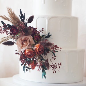May include: A white wedding cake with a drip icing design. The cake is decorated with a bouquet of dried flowers, including roses, pampas grass, and burgundy berries.