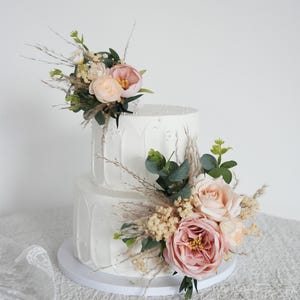 May include: Two-tiered white cake with textured frosting and pearl accents. The cake is decorated with a floral arrangement of pink and peach roses, greenery, and dried grasses, perfect for a wedding or celebration.