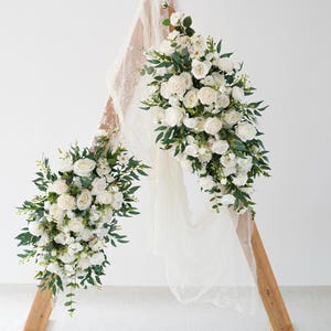 May include: A wooden wedding arch decorated with white roses and green foliage. The floral arrangements cascade down the arch, creating a romantic display. A sheer, cream-coloured fabric adds a soft touch to the design.