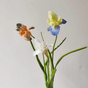 May include: A bouquet of three artificial iris flowers featuring one orange-brown bloom, one white bloom, and one bloom with yellow upper petals and blue lower petals, all attached to green stems with long, slender green leaves.