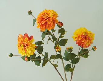 May include: A bouquet of three artificial orange and yellow dahlia flowers with green stems and leaves.