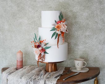 May include: A three-tiered white cake decorated with floral arrangements in shades of orange, peach, and burgundy. The cake sits on a wooden stand. A candle, teacup, and silverware are on a wooden table with a lace tablecloth.
