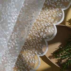 Puede incluir: Tela de encaje blanco con un delicado estampado floral. La tela está drapeada, revelando un borde festoneado. La imagen también incluye un cuenco marrón y ramitas verdes, creando una estética suave y natural.