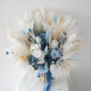 Op de afbeelding: Een bruidsboeket met gedroogd pampasgras, witte rozen en blauwe accentbloemen. Het boeket is vastgebonden met een blauw lint. Het arrangement heeft een zachte, luchtige textuur en een neutraal kleurenpalet met blauwe accenten.