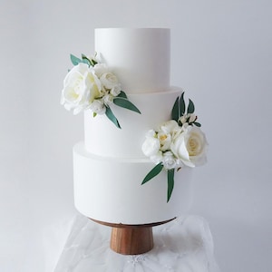May include: A three-tiered white cake decorated with white roses and green leaves. The cake sits on a wooden cake stand. The cake is a classic design, perfect for a wedding or other special occasion.