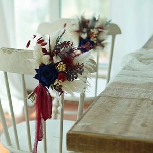 Puede incluir: Dos sillas de madera blanca decoradas con arreglos florales. Cada arreglo presenta una rosa azul marino, detalles en borgoña y elementos blancos plumosos. Una cinta burdeos cuelga del arreglo. Las sillas están cerca de una mesa de madera.