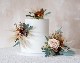 Op de afbeelding: Twee-laags witte bruidstaart versierd met gedroogde bloemen en groen. De taart is versierd met beige, perzikkleurige, bordeauxrode en oranje bloemen, evenals eucalyptusbladeren en gedroogde grassen.