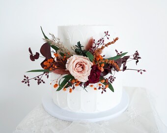 May include: A two-tiered white cake decorated with a floral arrangement. The arrangement includes a large pink rose, burgundy and orange flowers, green leaves, and dried grasses. The cake sits on a white cake stand.