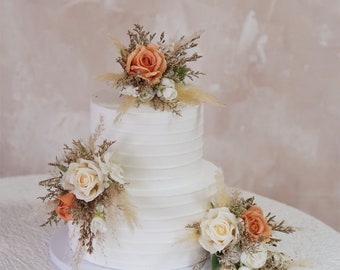 May include: A two-tiered white wedding cake decorated with dried flowers and greenery. The cake is adorned with peach and cream colored roses and beige dried grasses.