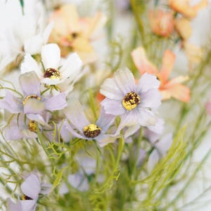 Puede incluir: Un ramo de delicadas flores artificiales de colores pastel. Las flores son una mezcla de blanco, rosa y lavanda, con centros amarillos. Las flores están dispuestas en un racimo, con algunos tallos que se extienden desde el centro.