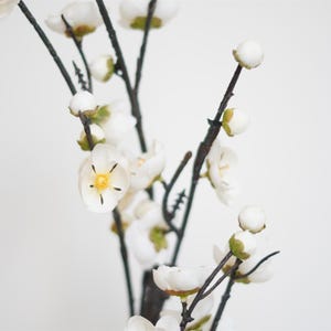 May include: A white artificial flower branch with delicate blossoms and buds. The branch has a natural, twig-like appearance and is perfect for adding a touch of spring to any space.