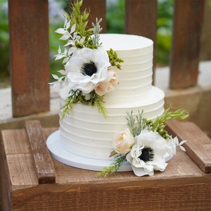Könnte beinhalten: Zweistöckige weiße Hochzeitstorte mit Buttercreme-Frosting und einem rustikalen Holzsockel. Die Torte ist mit weißen und pfirsichfarbenen Anemonen und Grün verziert.