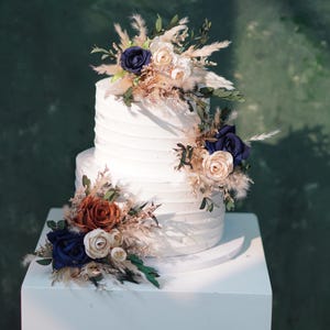 May include: A white two-tier wedding cake decorated with dried flowers and blue and brown roses. The cake is on a white cake stand.