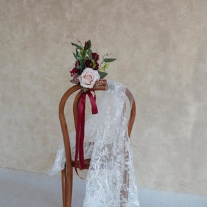 May include: A wooden chair adorned with a floral arrangement and a lace veil. The bouquet features a light pink rose, burgundy flowers, and greenery, tied with a burgundy ribbon. The white lace veil drapes over the chair.