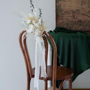 May include: A wooden chair adorned with a floral arrangement. The arrangement features white roses, pampas grass, and eucalyptus. White lace ribbons cascade down the chair. The chair is set against a neutral background with a dark green cloth.