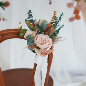 Op de afbeelding: Bloemdecoratie voor een stoel met een grote, lichtroze roos omringd door groen en gedroogde grassen. Een wit lint hangt aan de houten stoel. Ideaal voor bruiloften of speciale evenementen.