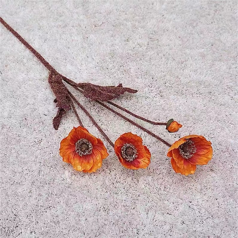 Puede incluir: Un ramo de cuatro flores de amapola artificiales de color naranja con tallos y hojas marrones. Las flores est&aacute;n dispuestas en un ramo y se encuentran sobre una superficie blanca y peluda.