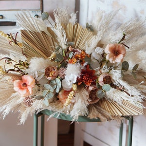 Pode incluir: Um arranjo floral seco com capim-dos-pampas, folhas de palmeira secas e flores de pêssego e laranja em estilo boho.