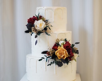 May include: A three-tiered white wedding cake with a drip icing design. The cake is decorated with red, white, and yellow flowers.