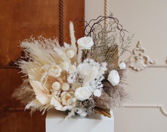 May include: A dried floral arrangement in a white vase. The arrangement features white pampas grass, dried palm leaves, white flowers, and brown branches.