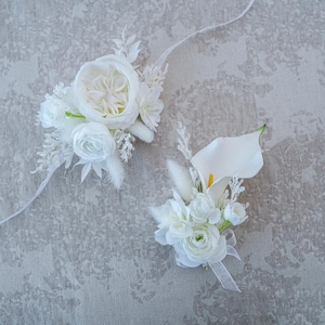 May include: Two white floral arrangements, one a wrist corsage and the other a boutonniere. The wrist corsage features a large rose and smaller white flowers. The boutonniere includes a calla lily and other white blossoms. Both are on a textured gray surface.