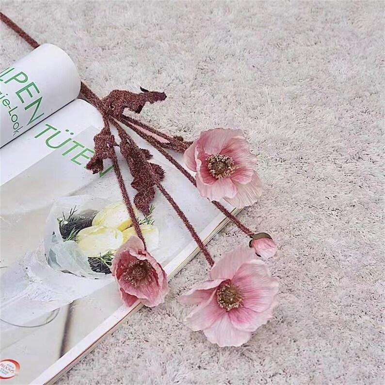 Puede incluir: Un ramo de cinco flores de amapola artificiales de color rosa con tallos y hojas marrones. Las flores est&aacute;n dispuestas sobre una superficie blanca y peluda.