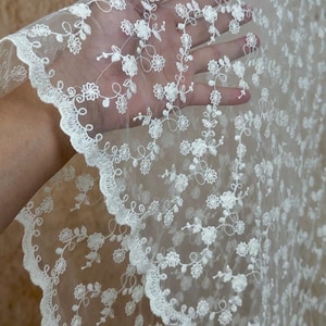May include: A sheer white lace fabric featuring an intricate floral embroidery pattern with delicate vines and small blossoms. The material has a scalloped edge with a decorative border, showcasing a fine mesh base with detailed white thread work.