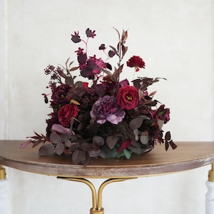 May include: A floral display of deep red and purple flowers and foliage. The arrangement is presented on a half-moon wooden table with gold-coloured metal legs and white accents. The flowers include roses and other blooms.