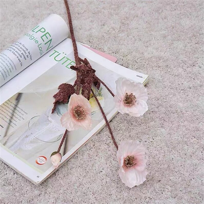 Puede incluir: Un ramo de tres flores de amapola artificiales de color rosa con tallos y hojas marrones. Las flores est&aacute;n dispuestas sobre un libro con ribete de encaje blanco.