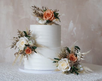 May include: Two-tiered white wedding cake decorated with dried flowers and pampas grass. The cake is frosted with a smooth white buttercream and has a rustic, elegant feel.