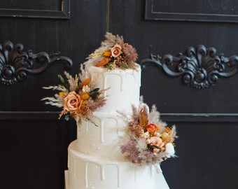 May include: A three-tiered white wedding cake decorated with dried flowers in shades of orange and brown. The cake has a drip effect with white icing.