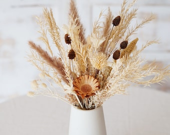 May include: A dried flower arrangement in a white vase. The bouquet features dried grasses, pampas grass, and a single brown flower.