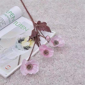 Puede incluir: Un ramo de flores de amapola de seda rosa con tallos y hojas marrones. Las flores est&aacute;n dispuestas sobre una superficie blanca y peluda.