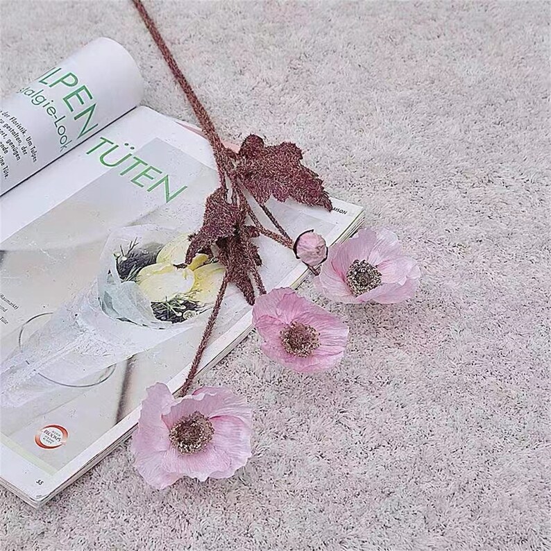 Puede incluir: Un ramo de flores de amapola de seda rosa con tallos y hojas marrones. Las flores est&aacute;n dispuestas sobre una superficie blanca y peluda.