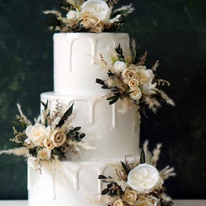 May include: A three-tiered white cake with a smooth finish and white icing drips cascading down each layer. The cake is decorated with clusters of cream-colored roses, dried beige grasses, and dark green foliage placed on each tier.