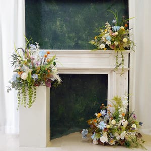 May include: A white painted fireplace mantel with three floral arrangements. The arrangements are made with a variety of flowers in shades of blue, yellow, and white. The flowers are arranged in a rustic style and are placed on top of the mantel and on the floor in front of it.