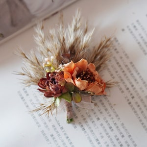 May include: A small bouquet of dried flowers with a peach and brown color scheme. The bouquet is tied with a brown ribbon and includes dried grasses and small white flowers.