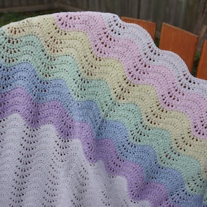 Peut inclure: Une couverture au crochet aux couleurs pastel avec un bord festonné. La couverture présente un motif de vagues dans les tons de blanc, lavande, bleu, vert et jaune. La couverture est drapée sur une chaise en bois.