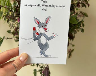 May include: A greeting card with a cartoon rabbit holding a red telephone, with the text "Yeah, so apparently Wednesday's hump day!" The rabbit is gray with a white belly and red accents. The card is held in front of a background of green plants.