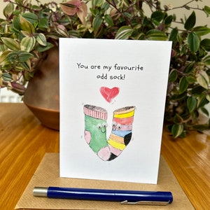May include: A greeting card with the text "You are my favourite odd sock!" features two cartoon socks with googly eyes and a red heart. A blue pen rests on a brown paper square in front of the card.