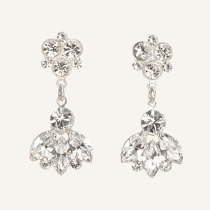 Peut inclure: Une paire de boucles d'oreilles pendantes argentées. Chaque boucle d'oreille présente un groupe de petits cristaux clairs ronds en haut, reliés à un groupe plus grand de cristaux en forme de goutte. Bijoux de soirée.