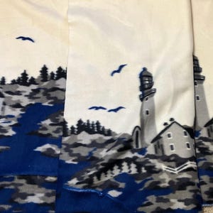 May include: White fabric with a coastal design featuring lighthouses, houses, and flying birds. The scene includes a blue river and gray and blue rocky terrain. The lighthouses are gray and white.