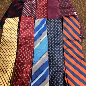 May include: A collection of ten neckties featuring various patterns and colors, including striped, paisley, and geometric designs in shades of red, blue, yellow, orange, and burgundy, arranged in two rows on a textured brown surface.