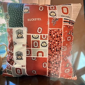 May include: A square throw pillow featuring a patchwork design of Ohio State Buckeyes themed fabric. The pattern includes various red, white, and grey panels with the text BUCKEYES, OHIO STATE, and the mascot Brutus Buckeye printed throughout.