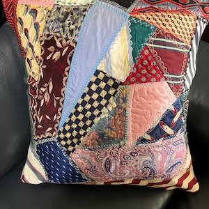 May include: A square throw pillow featuring a patchwork design composed of various fabric scraps in diverse colors and patterns, including paisley, checkered, floral, and striped motifs, stitched together with visible decorative embroidery.